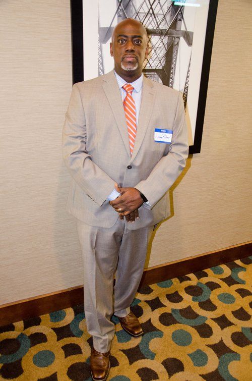 Man in tan suit, orange tie, standing near a wall with art and patterned carpet.