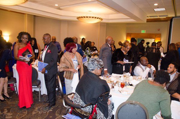 Guests mingling in a brightly lit banquet hall. Several seated at round tables; some standing, chatting, holding programs.