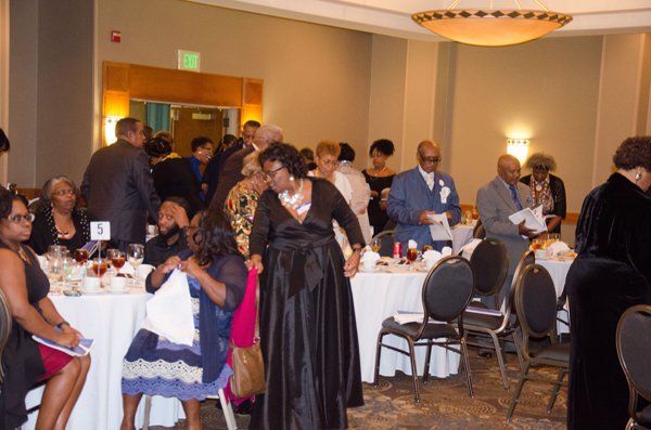 People gathered at tables in a banquet hall, some standing, some seated. Tables are set with white linens.