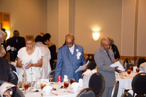People at an event seated at round tables. Some are holding papers, dressed in suits and dresses.
