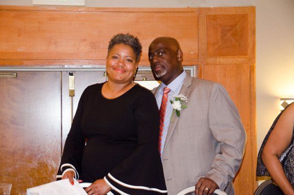 A smiling couple poses indoors; the woman in black dress, the man in a tan suit with a red tie.
