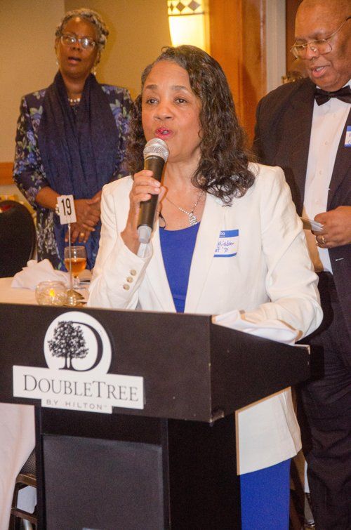 Woman speaking at a DoubleTree podium, wearing a white blazer, blue dress. Others are in the background.