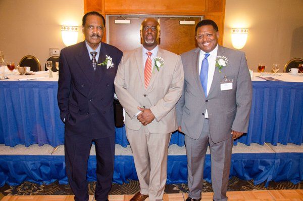 Three men in suits stand in front of a blue-skirted table.