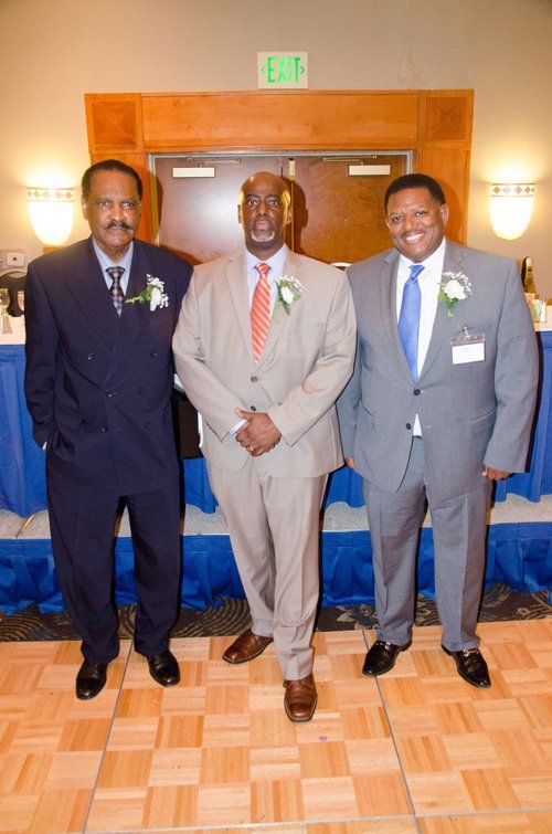 Three men in suits stand indoors near a blue draped table.