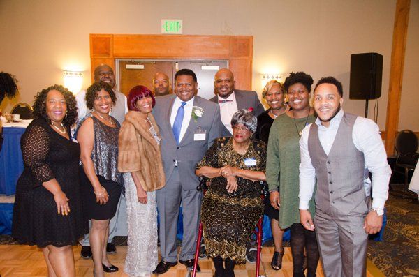 Group of people smiling for a photo indoors, some wearing formal attire, near a doorway, with a wheelchair.
