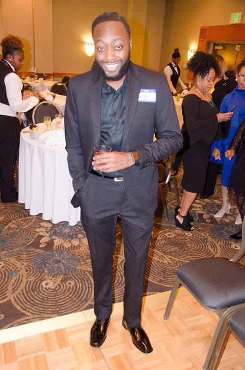 Man in dark suit holding a drink, smiling, standing in a banquet hall.