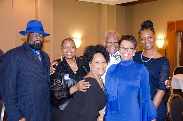 Group of six people smiling at a gathering; some wear blue. Indoors, warm lighting.