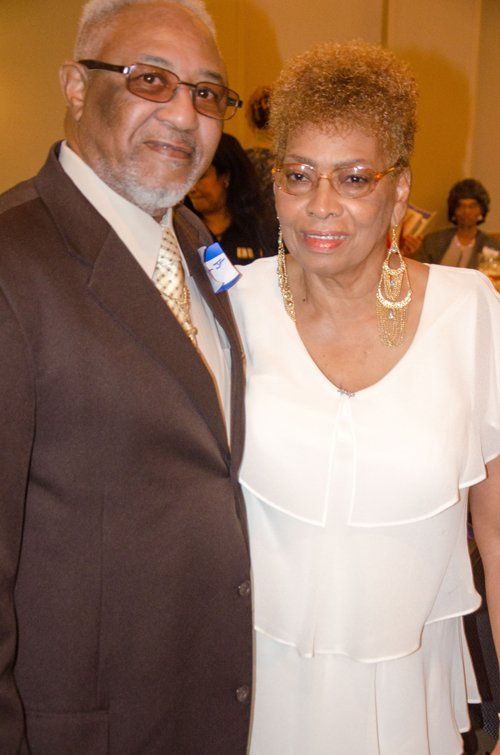 Couple in formal attire smiling indoors. Man in brown suit, woman in white dress, gold jewelry.
