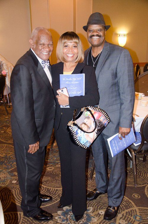 Three people posing together at an event. Woman in black suit holds a purse and document. Men in suits smile.