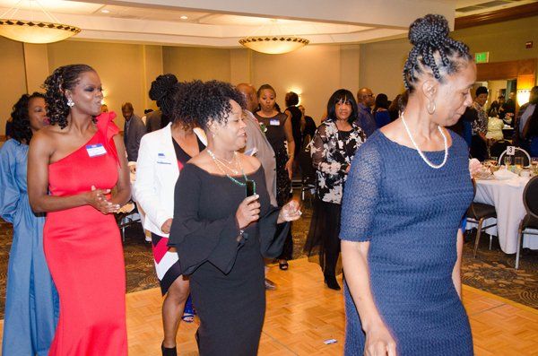 People dancing at an event in a ballroom. Woman in red dress, black dress with mic. Others look on.