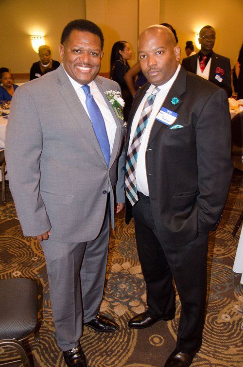 Two men in suits pose indoors at an event, smiling. One wears a light suit, the other a dark suit.