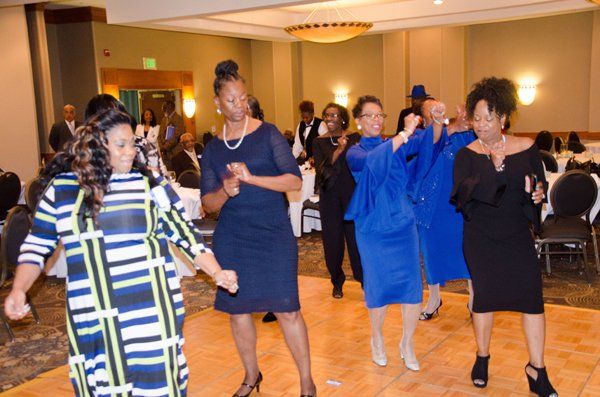 People dancing on a wooden dance floor in a ballroom; multiple in blue and black dresses.