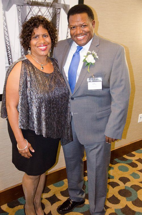 Couple smiling and posing together. The man wears a suit with a blue tie, and the woman wears a sequined top and skirt.