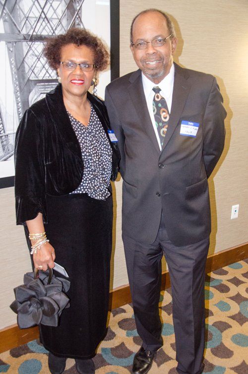 Woman in black ensemble stands next to man in a suit. They are indoors, smiling.