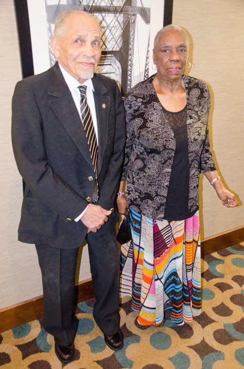 An older man and woman standing together indoors; man in suit, woman in patterned dress.