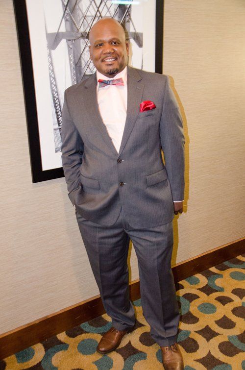 Man in gray suit with pink bow tie and pocket square, brown shoes, smiling, in a hallway.