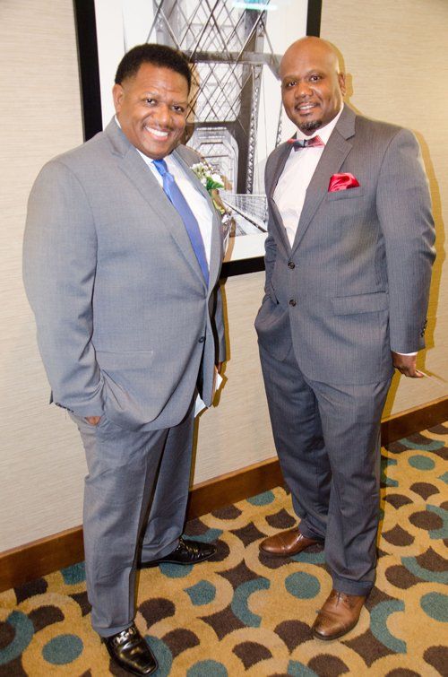 Two men in suits pose for a photo indoors, smiling. One in blue tie, the other in a bow tie and pocket square.
