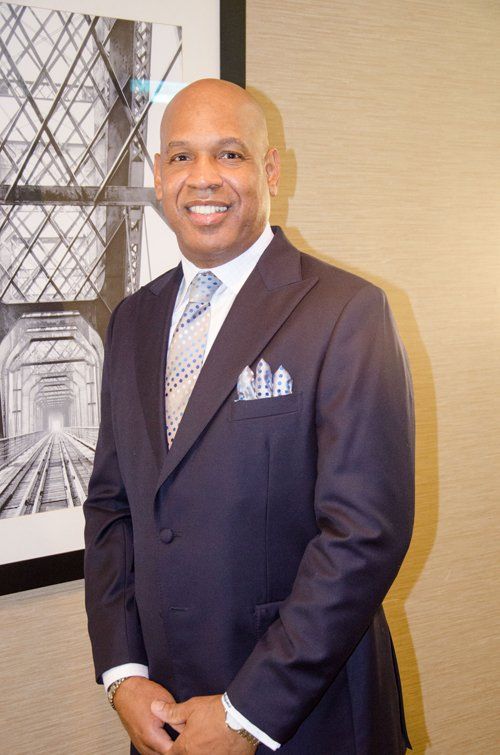 Man in navy suit, smiling, in front of abstract photo. Pocket square visible.