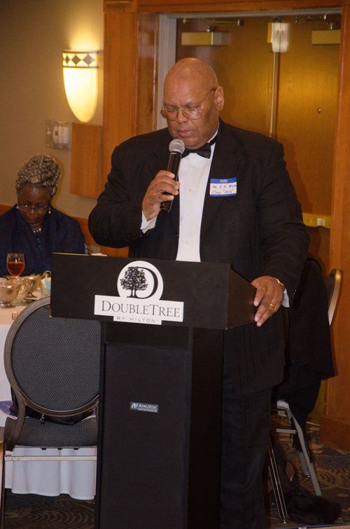 Man speaking into a microphone at a podium with a DoubleTree logo. Wearing a tuxedo, in a banquet hall.