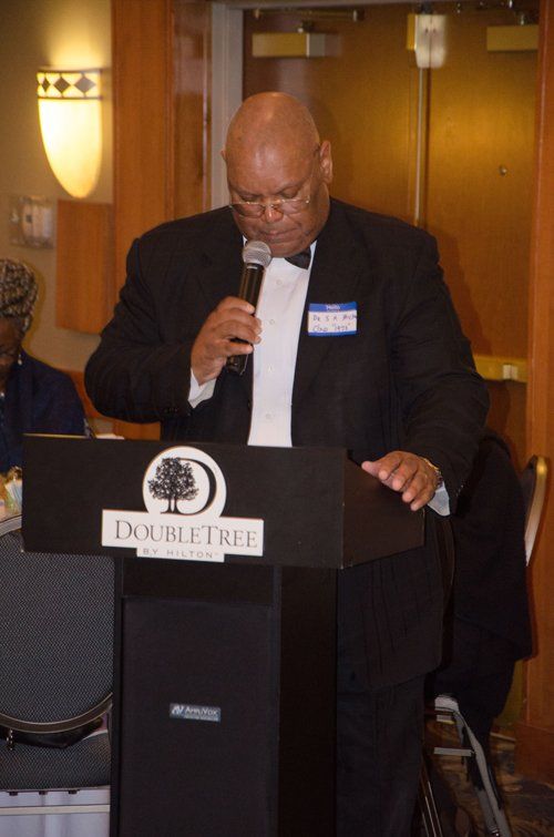 Man speaking at a DoubleTree podium, holding a microphone. He wears a black suit and bow tie.