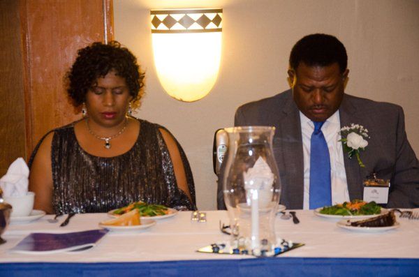 Man and woman at a table with food, eyes closed in prayer or reflection. Formal attire.