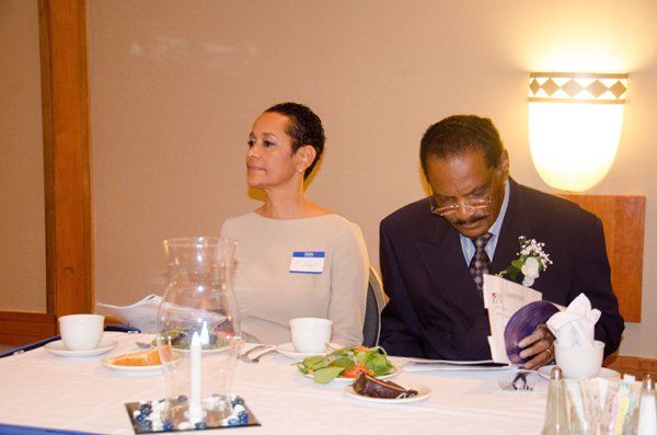 Woman and man at a table. The man is looking down, the woman is looking up. Light tan walls and a lamp.