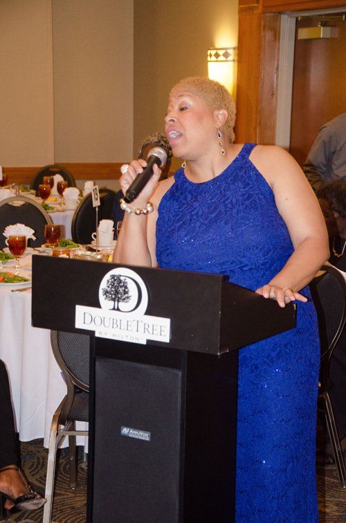 Woman in blue dress speaks into a microphone at a DoubleTree podium. Restaurant setting.