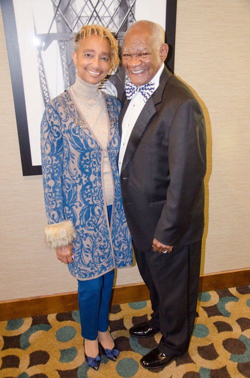 Woman in patterned blue coat, man in tuxedo, smiling, posing together indoors.