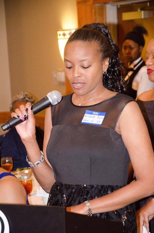 Woman speaking into a microphone at a formal event, wearing a black dress.