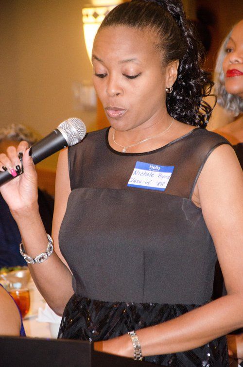 Woman speaking into microphone at event, wearing black dress, name tag visible.
