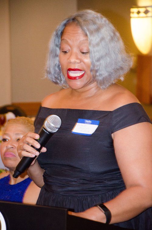 Woman with silver hair speaking into a microphone at an event, wearing a black off-the-shoulder dress.