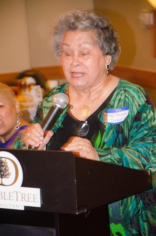Woman speaking into microphone at a podium, wearing green patterned top, DoubleTree hotel logo visible.
