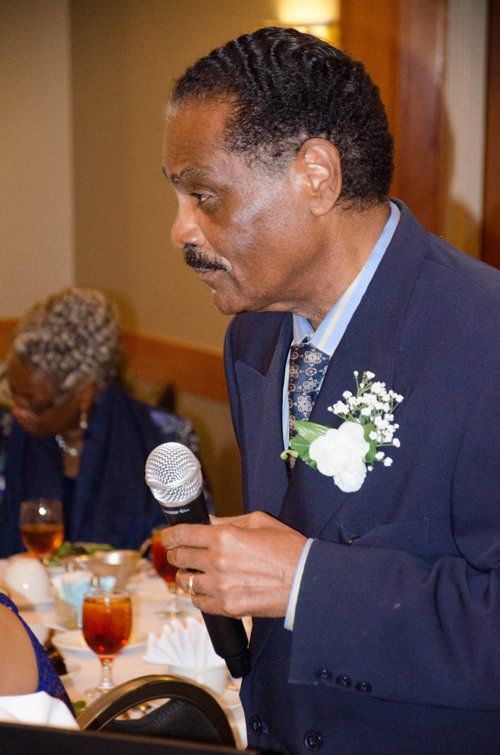 Man in a blue suit speaks into a microphone at a table. A woman is in the background.