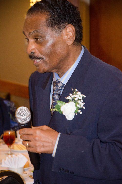 Man in blue suit speaks into a microphone at an event, with white flower pinned on his lapel.