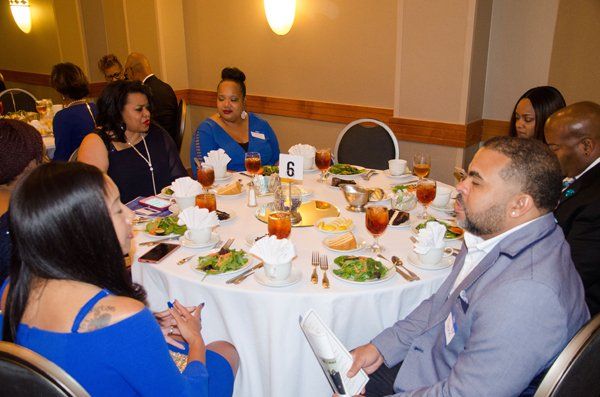 People seated around a round table set for a formal event. Food and drinks visible.