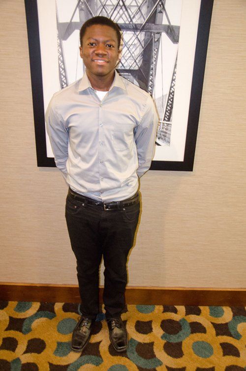 Man in grey shirt and black pants stands in front of a picture. He has his hands behind his back and is smiling.