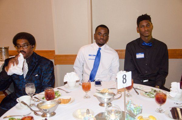 Three men seated at a formal dinner table, one wiping his mouth, another with a blue tie, the third with a blue bow tie.