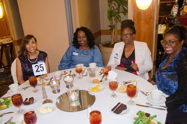 Four people sit at a round table, smiling. There's food and drinks, a mirror centerpiece.