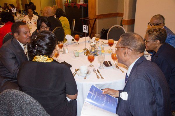 People seated around a white tablecloth table at a formal event, conversing. Some have drinks and menus.