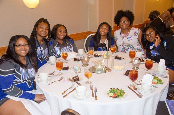Group of people at a round table, smiling, some wearing blue and grey shirts. Plates of food, drinks.