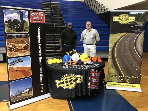 Two men at a Lehman Roads booth with safety equipment and company banners in a gymnasium.