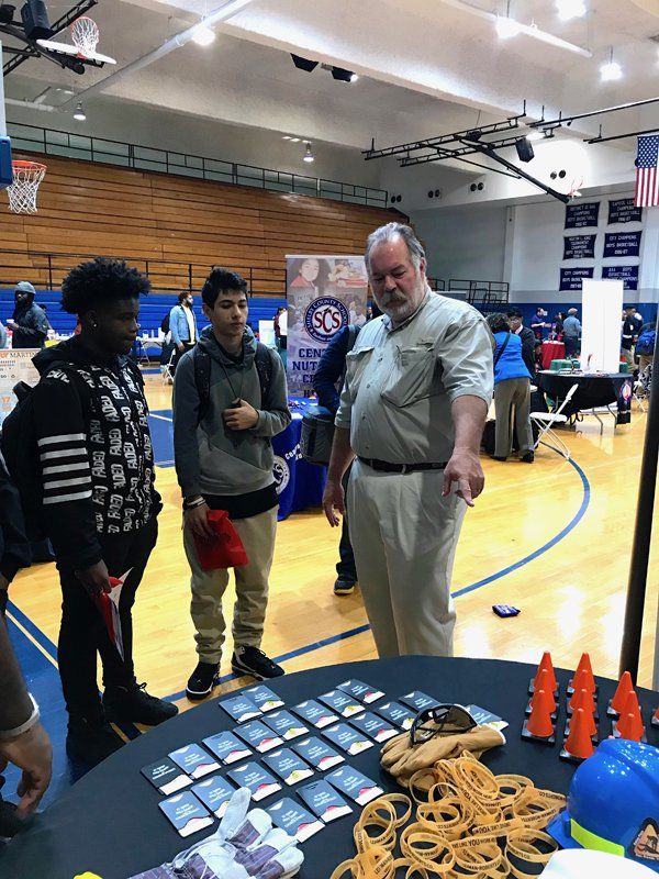 Man points at table with students in a gym. Table displays cards, rubber bands, and cones.