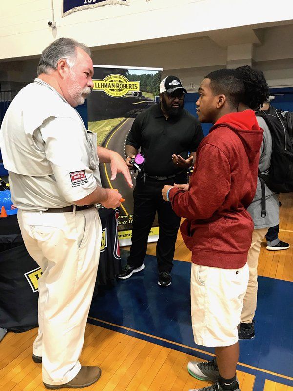 Man in tan shirt gestures while talking to a teen in a red hoodie; another man stands nearby.