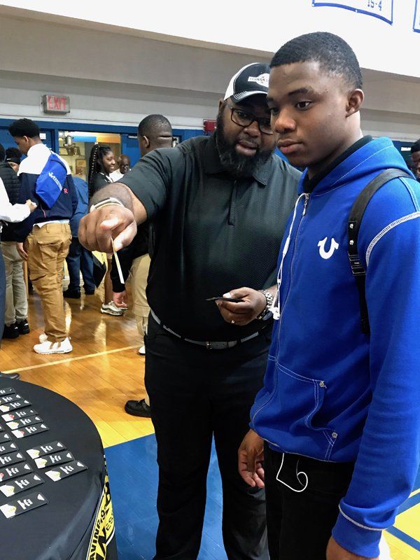 Man points at items on a table for a student wearing a blue hoodie in a school gymnasium.