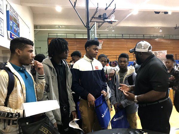Group of teens listening to a man in a gymnasium, possibly at a school event.