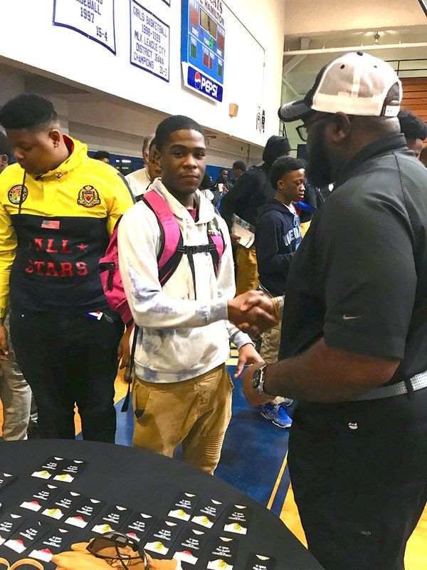 Man shaking hands with another man at a table with small items. People stand nearby in a gym.