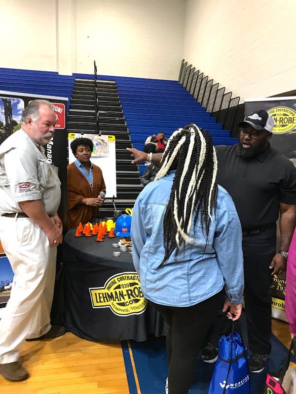 People at a booth promoting Levman-Robar. Man points; another looks at products. Blue gym bleachers in background.
