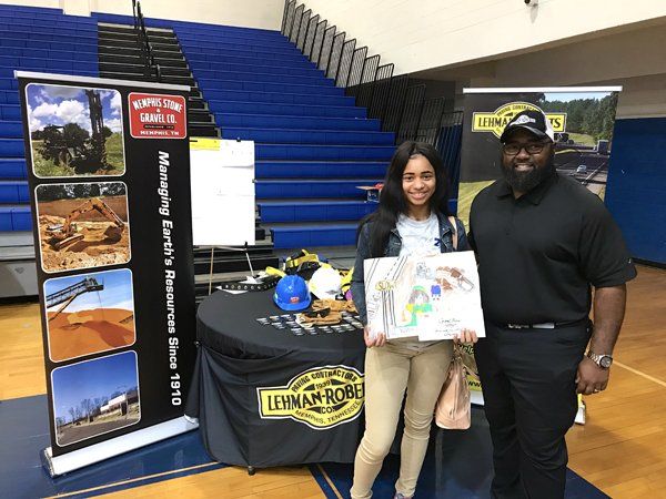 Woman and man stand at a table with construction company banners. The woman holds a drawing, smiles.