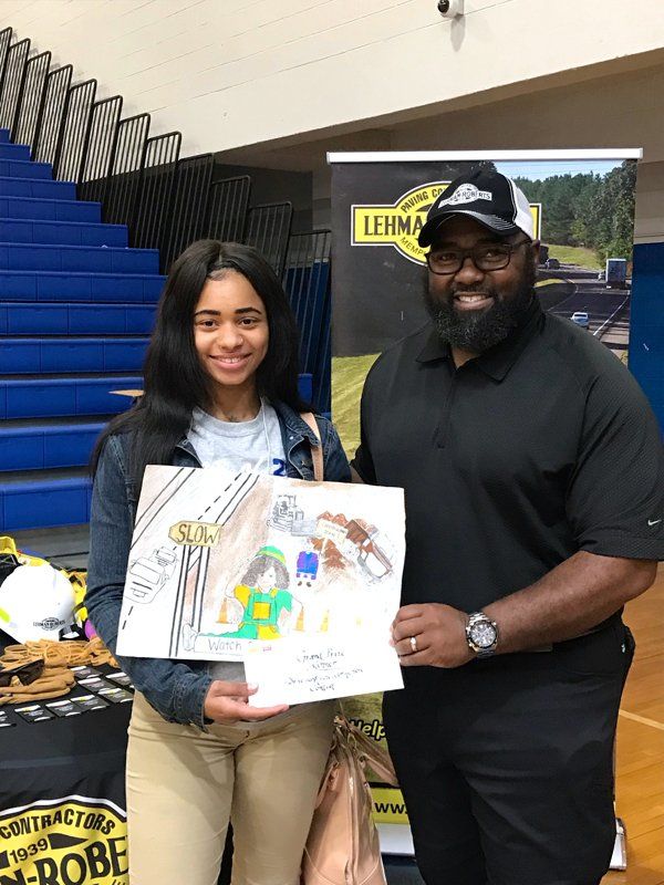 Teenage girl and man hold up a drawing, smiling. Backed by Lehman Contractors signage in gym.