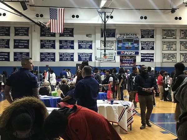 People at a school event in a gymnasium, tables set up. American flag and basketball hoop visible.